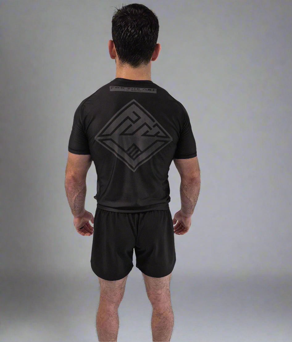 Person wearing a black t-shirt and shorts with a logo on the back, standing against a white curtain.